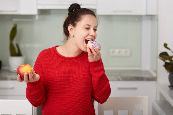 Pregnant woman in kitchen holding apple in hand and eating cake. Young attractive female decides to eat unhealthy food, looks happy, dressed casually, has hairstyle bun, posing in white kitchen. - Photo, Image