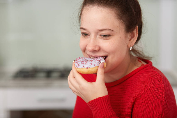 Close up portrait of young happy woman eating cake while sitting at table in kitchen, lady wearing red casual sweater, has happy facial expression, looking down. Unhealthy eating and lifestyle concept - Fotografie, Obrázek