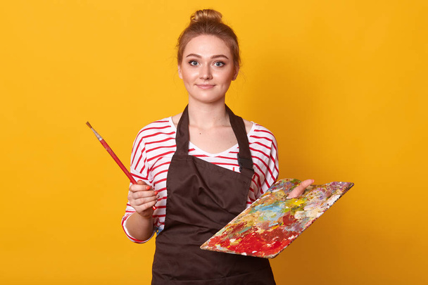 Surprised emotional blond haired female posing with bunch on her head, having impressed facial expression, holding paint equipment for her hobby, isolated over bright yellow background in studio. - Foto, Bild