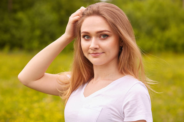Close up portrait of young beautiful blonde woman with staright hair, posing in meadow in sunny summer or spring day, looking directly at camera, keeps hand on head, dressed casual white t shirt. - 写真・画像