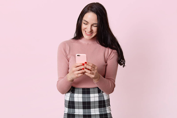 Portrait of happy smiling young woman in casual shirt and checkered skirt, using mobile phone, typing messages or checking social networks. People sincere emotions, lifestyle concept, posing in studio - Fotografie, Obrázek