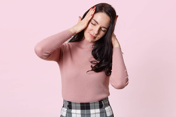 Studio shot of attractive young woman with terrible headache, keeps both hands on head, needs to take medicine, brunette female in rose shirt and checkered skirt, isolated over pink background. - Zdjęcie, obraz