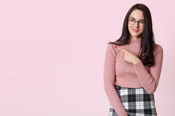 Indoor studio shot of pleasant brunette with sincere smile on her face, showing with forefinger aside, making gesture, wearing sweater, formal skirt and trendy specs. Copyspace for advertisement. - Fotó, kép