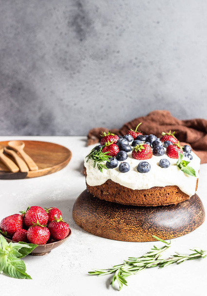 Wholegrain cake with zucchini, carrot and walnuts decorated with whipped cream, strawberries, blueberries, mint and rosemary on a wooden plate.  - Фото, изображение