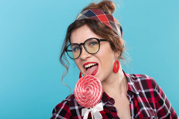 Close-up portrait of a charming young pin-up woman in a plaid shirt and glasses is licking a big lollipop against a blue background. Concept of love for sweets and desserts. Copyspace. - Foto, Imagem