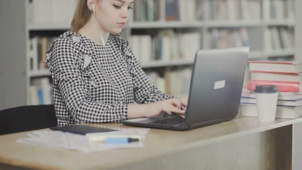 Piękna studentka w słuchawkach siedząca przy biurku w bibliotece pracująca skoncentrowana na projekcie szkolnym. - Materiał filmowy, wideo