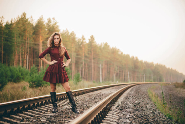 Beautiful dreamy girl with curly natural hair enjoy nature in forest on railway. Dreamer lady in burgundy dress walk on railroad. Inspired girl on rails at dawn. Sun in hair in autumn. Good mood. - Photo, Image