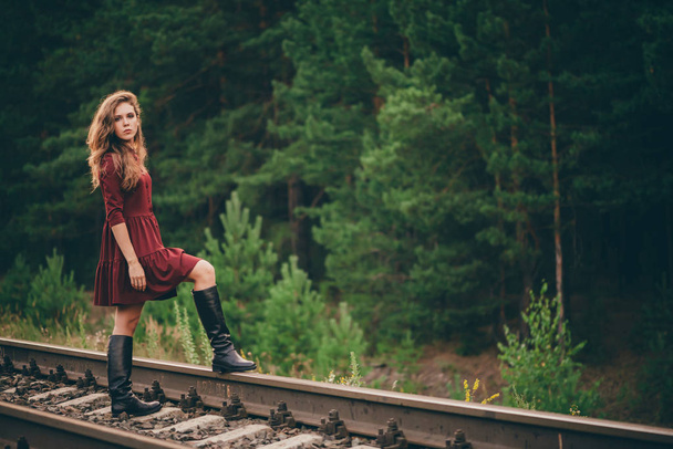 Beautiful sad thoughtful girl with curly natural hair on nature in forest on railway. Dreamer lady in burgundy dress walk on railroad. Depressed girl on rails at dawn. Sun in hair in autumn. Bad mood. - Фото, изображение