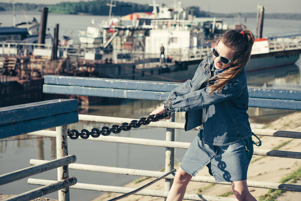 Adolescente amusante en robe de denim bleu marche dans la gare fluviale. Jeune enfant joyeux pisser dans le port de la rivière. Fille en lunettes de soleil tenir sur la chaîne
. - Photo, image