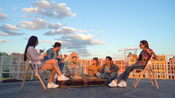 Skupina šesti přátel sedícího na střeše. Mluví, usmívají se a jedí pizzu. Čas západu slunce. Střešní strana. - Záběry, video