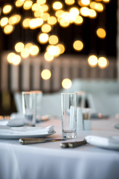 Ambiente de mesa em um restaurante. Copo de vidro limpo perto. Detalhes da mesa do banquete. No borrão de fundo estão queimando guirlandas com lâmpadas - Foto, Imagem