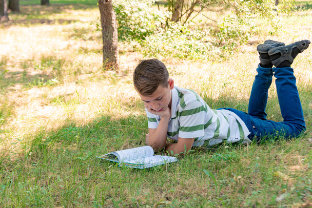 O menino está no parque na grama e está estudando na natureza, educação e ciência, o livro está deitado na grama
. - Foto, Imagem