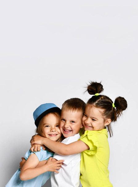 Studio portrait of children on a light background: full body shot of three children in bright clothes, two girls and one boy. Triplets, brother and sisters. hugging on camera. Family ties, friendship - Φωτογραφία, εικόνα