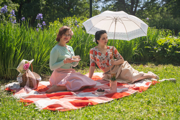 twee mooie pin-up dames met leuke picknick in het stadspark op een zonnige dag samen. meisjes vrienden genieten van hete zomer weer. mooie vrouwtjes in retro vintage stijl zijn ontspannen op het gras. - Foto, afbeelding