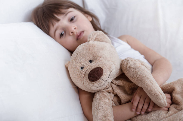 Милая маленькая девочка улыбается, лежа в уютной белой кровати
 - Фото, изображение