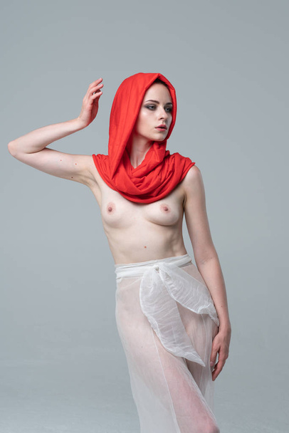 mladá krásná dívka pózuje nahá ve studiu, stojí v červené šále a bílé kalhoty - Fotografie, Obrázek
