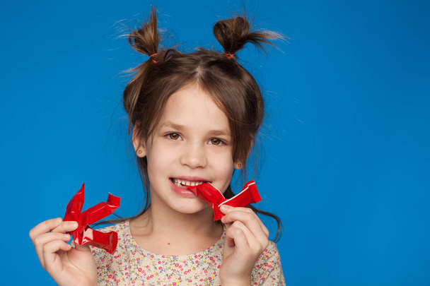 Portrait of a beautiful girl of five years old with an interesting hairstyle and with candies in her hands on a blue background - Fotoğraf, Görsel