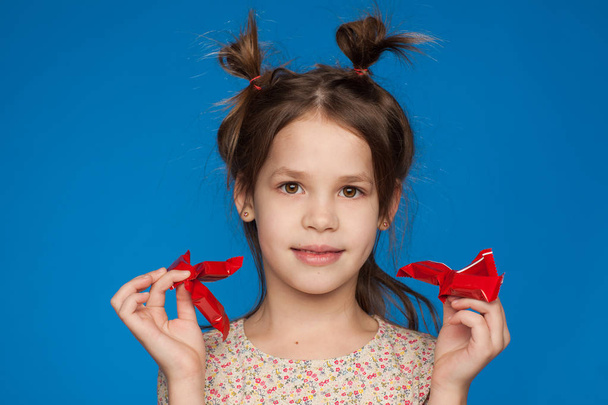 Portrait of a beautiful girl of five years old with an interesting hairstyle and with candies in her hands on a blue background - Zdjęcie, obraz