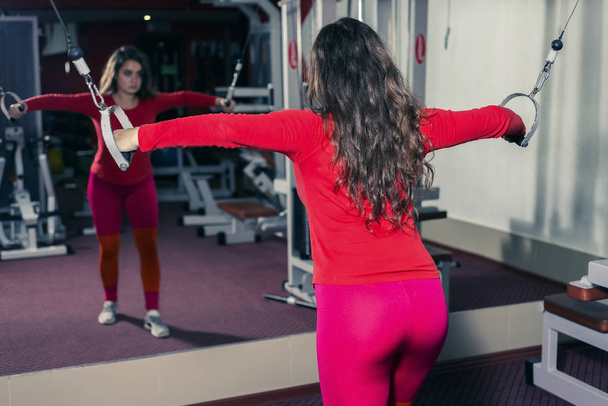 αθλητικό κορίτσι που ασχολούνται με το γυμναστήριο στον εξομοιωτή. άθλησης και την άρση βαρών. γυναίκα κοιτάζει μέσα από τον καθρέφτη - Φωτογραφία, εικόνα