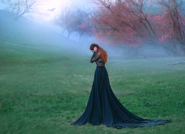 dark witch made terrible mistake, sad lady in long black dress with long tail and lace sleeves, girl with bright red orange hair pressed hands to head, autumn landscape, mysterious white haze - Фото, изображение