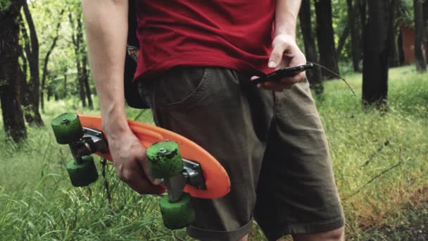 Mladý kavkazský hipster muž s skateboardem pobyt v parku a textové zprávy na mobilním telefonu - Záběry, video