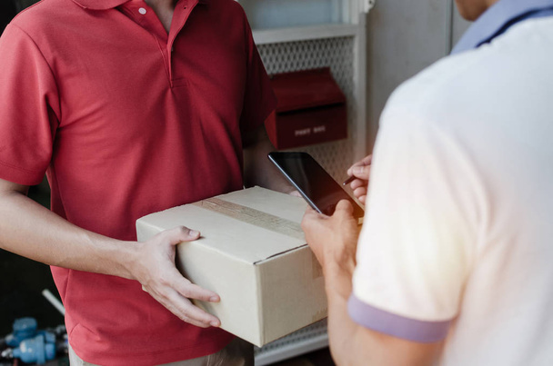 young man customer appending signature in digital mobile phone receiving parcel post box from courier with home delivery service man smiling face in red uniform at home, express delivery concept - Photo, image