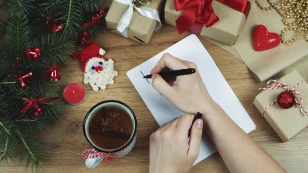Γυναίκα χέρια γράφοντας το νέο έτος για να κάνει λίστα σε λευκό φύλλο χαρτιού, σχεδιάζει το μέλλον της. Χριστουγεννιάτικη διακόσμηση με κλαδιά ελάτης, δώρα. Επίπεδη. Στο πάνω μέρος. Mockup, αντιγράψτε χώρο για το κείμενό σας. - Πλάνα, βίντεο