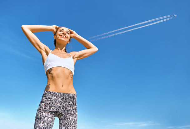 Щаслива усміхнена спортивна жінка з татуйованим тілом та татуюванням позує на блакитне небо з літаючими стежками
. - Фото, зображення