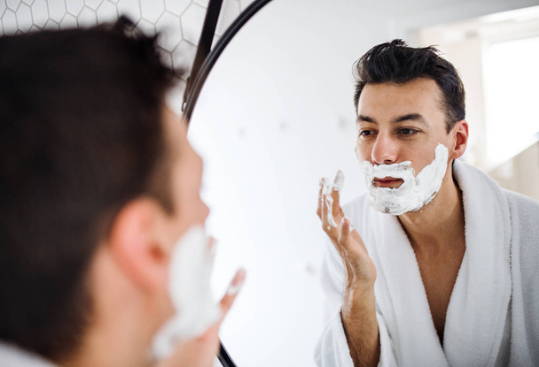 Young man with shaving foam in the bathroom in the morning, daily routine. - 写真・画像