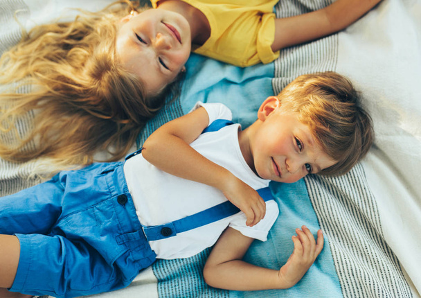 Над видом детей, лежащих на одеяле у травы. Счастливый мальчик и симпатичная девочка наслаждаются летом в парке. Очаровательные дети улыбаются и веселятся на солнечном свете на открытом воздухе. Детство
 - Фото, изображение