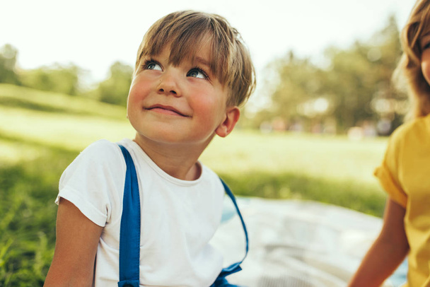 Улыбающийся мальчик, сидящий на одеяле на фоне природы. Счастливый ребенок наслаждается летом со своей сестрой в парке. Красивый ребенок улыбается и веселится на солнечном свете на открытом воздухе. Детство
 - Фото, изображение