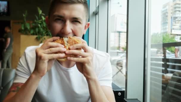 Νεαρός όμορφος τύπος τρώει χάμπουργκερ σε ένα εστιατόριο, φαστφουντάδικο - Πλάνα, βίντεο