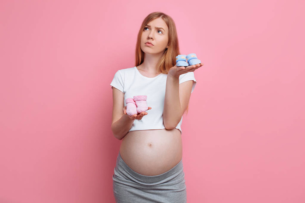 Beautiful pensive pregnant woman holding blue and pink baby booties, posing in the studio on a pink background - Фото, зображення