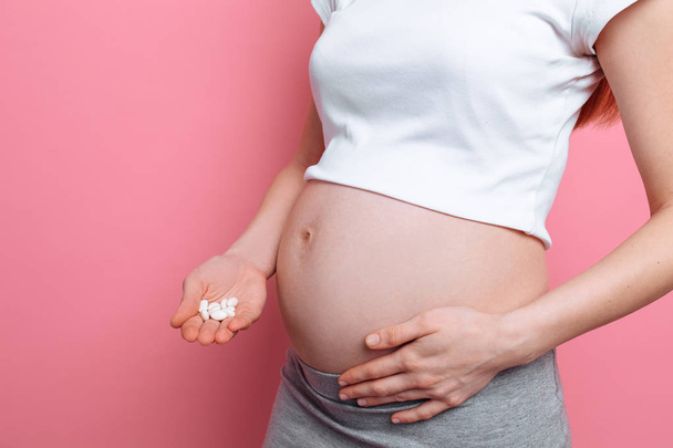 Estómago de una mujer embarazada sosteniendo píldoras blancas redondas para el cuidado de la salud, primer plano del cuerpo humano sin rostro
 - Foto, Imagen