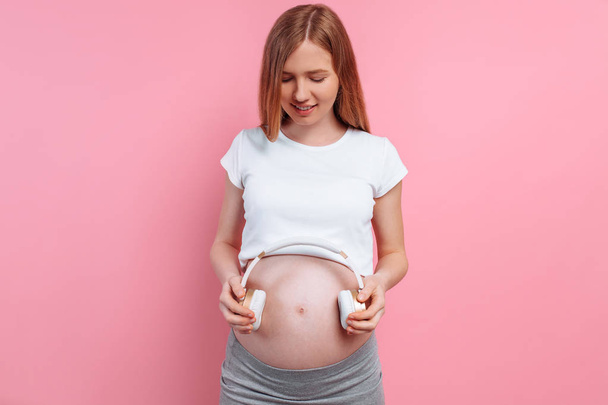 Attractive pregnant woman holding headphones on her belly over a pink background - Foto, imagen