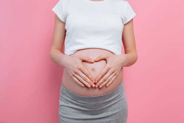 Μια έγκυο γυναίκα που κρατάει τα χέρια της σε σχήμα καρδιάς στην κοιλιά της. Έγκυος κοιλιά με δάχτυλα. Σχήμα καρδιάς σε ροζ φόντο. - Φωτογραφία, εικόνα