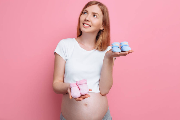 Beautiful pensive pregnant woman holding blue and pink baby booties, posing in the studio on a pink background - Foto, Imagen