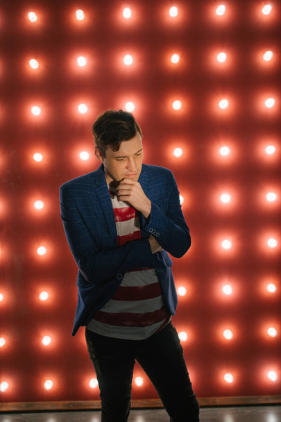 Young male standing against illuminated wall. Trendy man looking at camera on red background with light bulbs - Фото, зображення