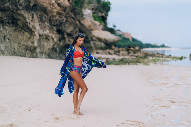 Beautiful sexy girl in swimwear walking on sandy beach with rock on background - Foto, afbeelding