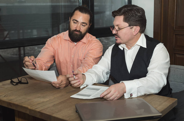 Dos hombres de negocios discutiendo documentos financieros mientras están sentados a la mesa en la oficina
. - Foto, imagen