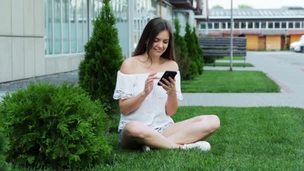 Young happy woman uses the phone while sitting on the lawn near the hotel prints a message - 映像、動画