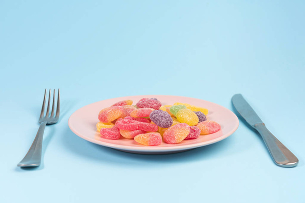 Fork, knife and pink plate with gummy candies on blue background - Fotoğraf, Görsel