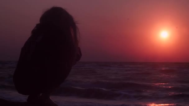 schönes Mädchen beobachtet Sonnenuntergang am Meer dunkle Farbe sitzt auf dem Stein - Filmmaterial, Video