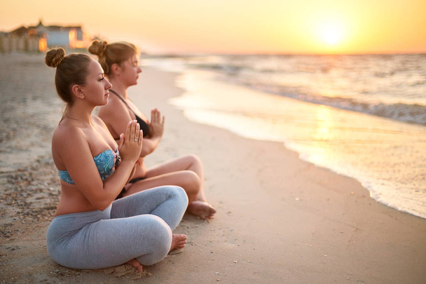 Két hiteles nő csinál csoport jóga meditáció a tengerparton napkelte. Lányok pihentető lótusz jelentenek Asana a tengeren parton. Body pozitív nőstények gyakorló Padmasana az óceán közelében surf reggel. - Fotó, kép