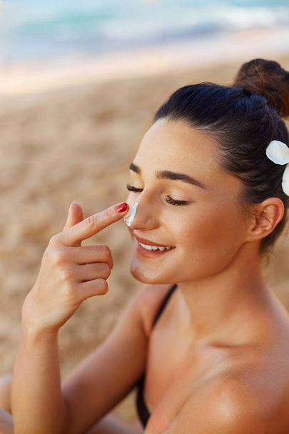 Красивая счастливая милая женщина наносит крем для загара на нос на пляже. Защита тела от солнца. Крем от солнца. Бикини женщина мажет увлажняющий лосьон на кожу. Suntan
 - Фото, изображение
