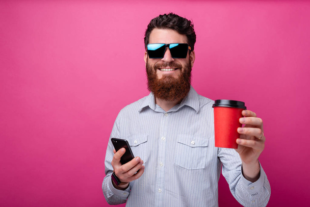 Young cheerful bearded man holding a take away cup and his phone on pink background. - Photo, Image