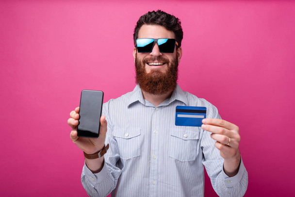vousatý muž, který nosí sluneční brýle, ukazuje na kameru jeho telefon s prázdnou obrazovkou a kreditní kartou na růžovém pozadí. - Fotografie, Obrázek