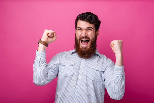 Молодий бородатий чоловік робить жест переможця на рожевому фоні
.  - Фото, зображення