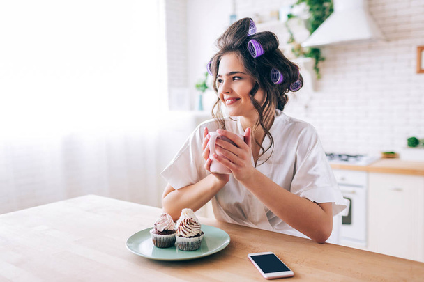 Beautiful young female housewife in kitchen. Holding cup and look to side. Smiling alone. Pancakes and phone on table. Carefree young woman living beatufiul rich life. No work. - Photo, image