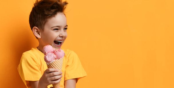 Baby boy kid hold strawberry pink ice-cream in waffle cone happy laughing looking at the corner on yellow - Fotó, kép